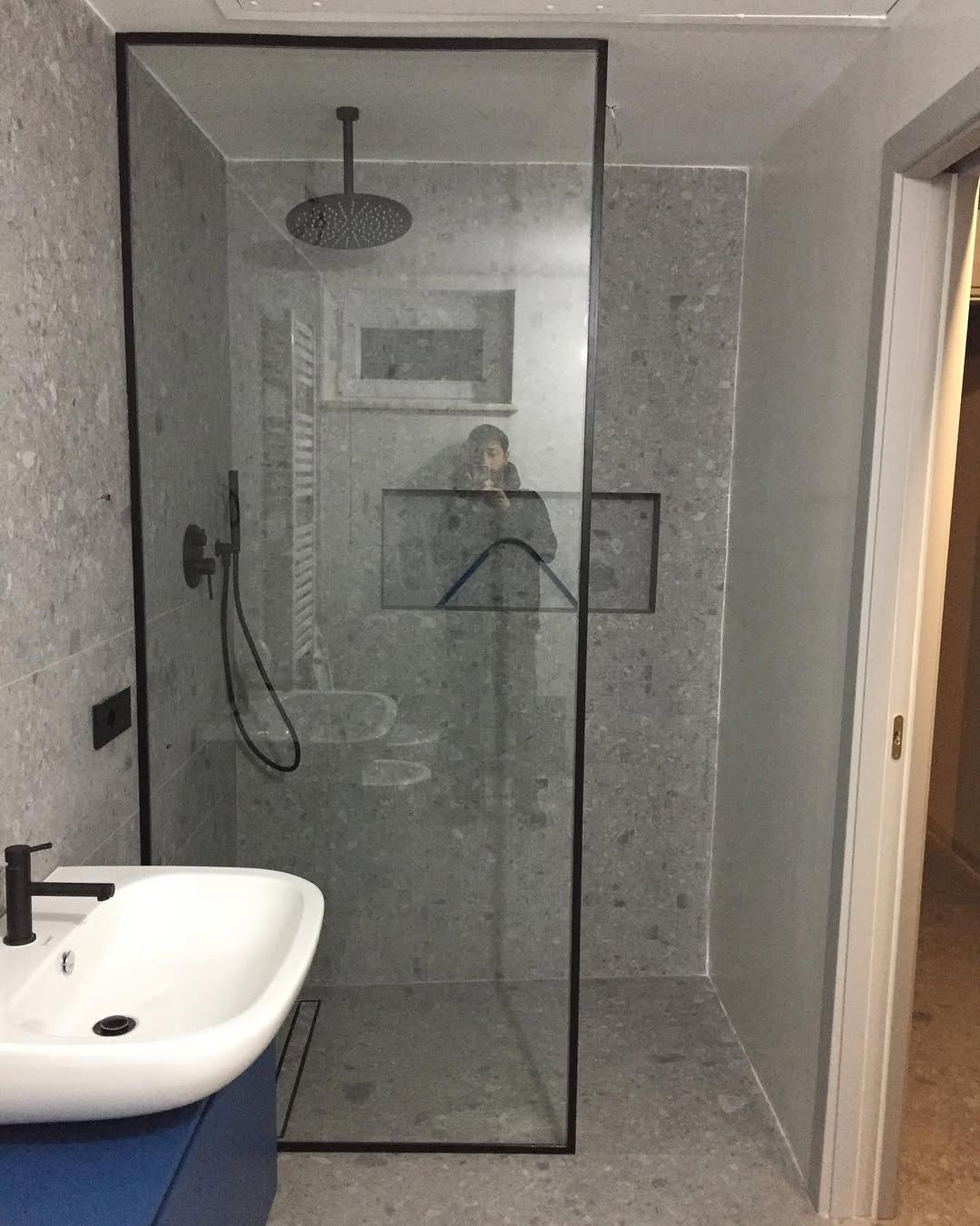 Bagno moderno con cabina doccia, parete divisoria in vetro, lavabo bianco e pareti rivestite con piastrelle grigie.
