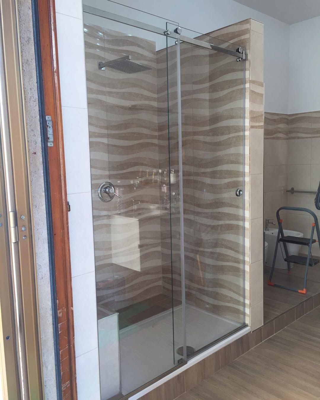 Box doccia in vetro in un bagno piastrellato con pareti a righe beige e marroni e pavimento effetto legno.