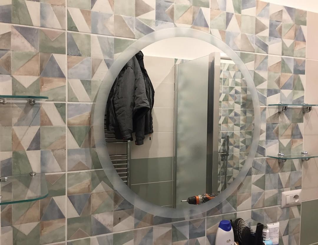 Specchio rotondo da bagno su una parete piastrellata con motivi decorativi, che riflette un cappotto appeso e la zona doccia.