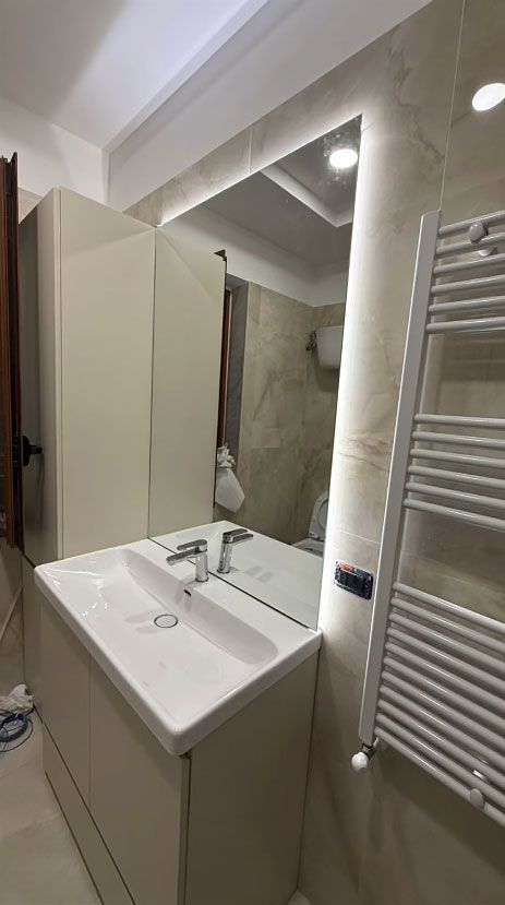 Lavabo e specchio del bagno con mobiletto bianco, parete piastrellata e scaldasalviette sulla destra.