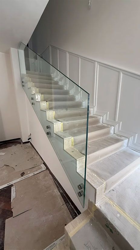 Scala bianca con ringhiera in vetro all'interno di un'abitazione non ancora ultimata, con detriti di costruzione sul pavimento.