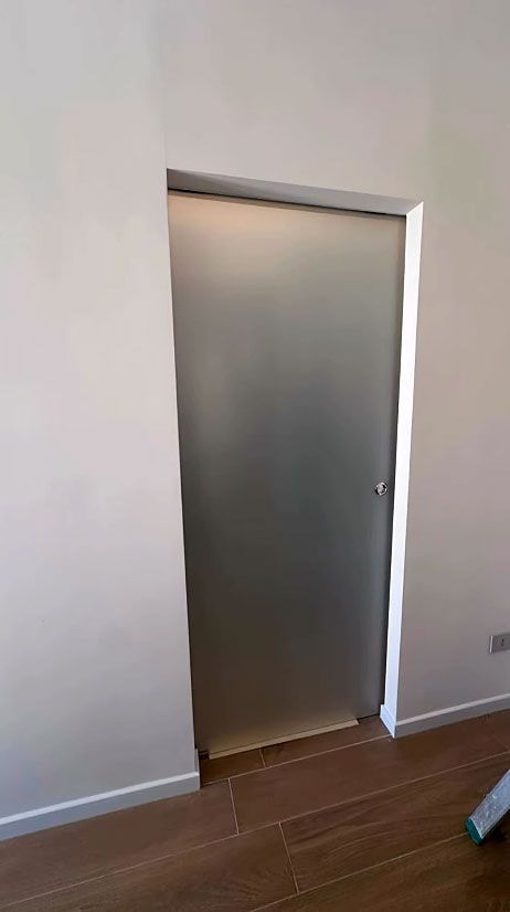 Stretta porta grigia in una stanza con pareti bianche e pavimento in legno, vista da una leggera angolazione.