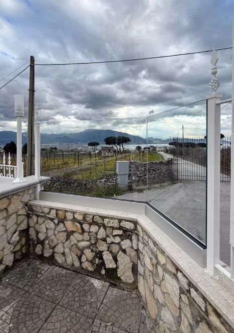 Muro di pietra e recinzione bianca che si affacciano su un paesaggio nuvoloso e montagne in lontananza.