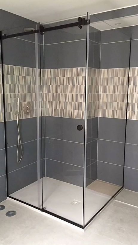 Box doccia angolare in vetro con pareti rivestite di piastrelle scure e piatto doccia bianco.