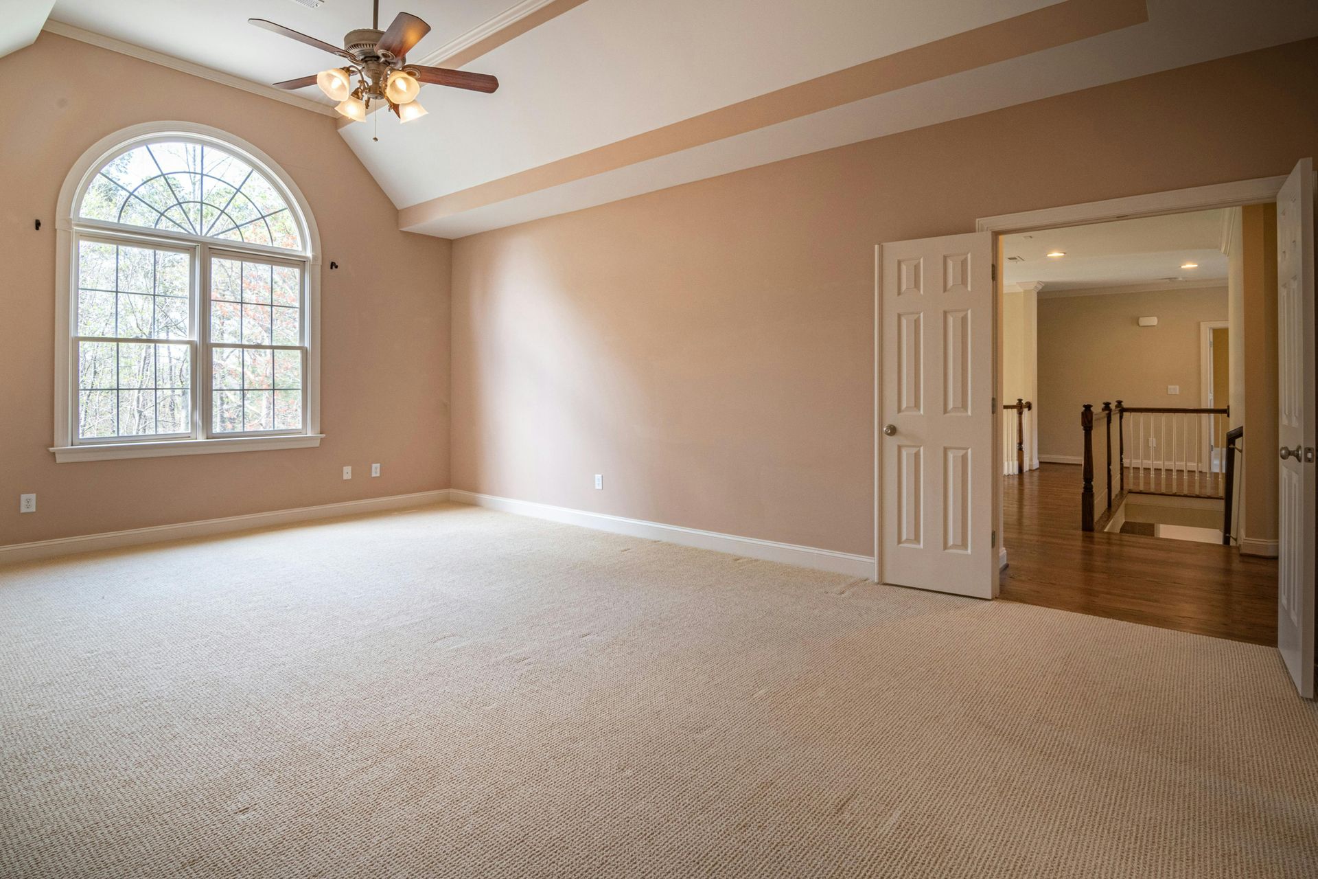 Empty beige bedroom with vaulted ceiling, arched window, ceiling fan, and open double doors to hallway