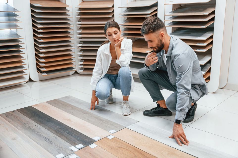Two people crouch on a showroom floor, examining different wood-look flooring samples laid out for comparison.
