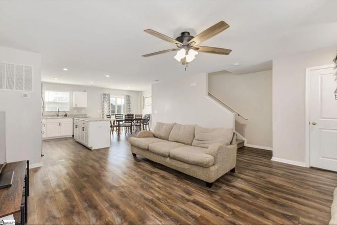 An open-concept living area with wood floors, a beige couch, a ceiling fan, and a kitchen visible in the background.