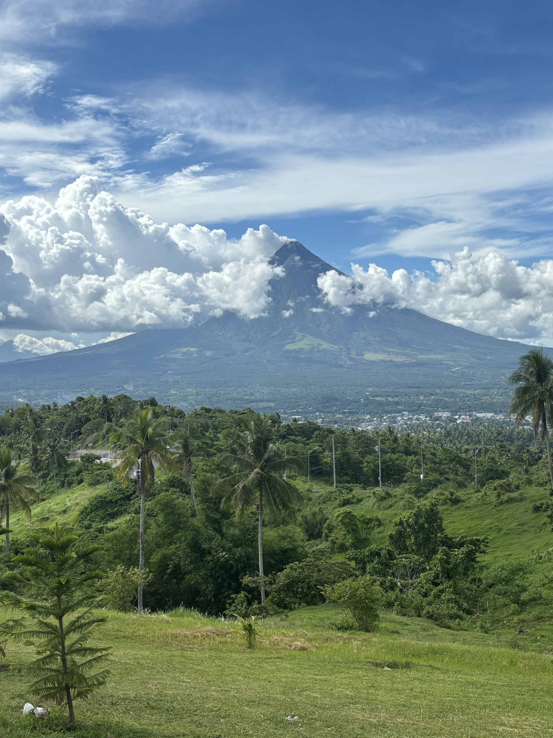 Lush green landscape with a volcano partially covered in white clouds against a bright blue sky.