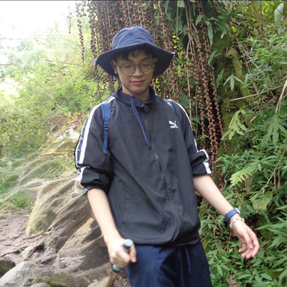 Person in blue hat and jacket holds walking stick on a trail with greenery.