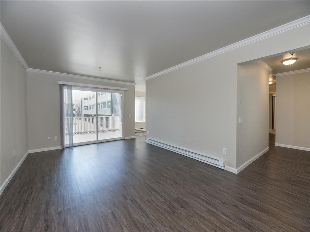Living room with sliding glass door to balcony, light walls, and dark laminate flooring at Tower apartments in Alameda, CA.