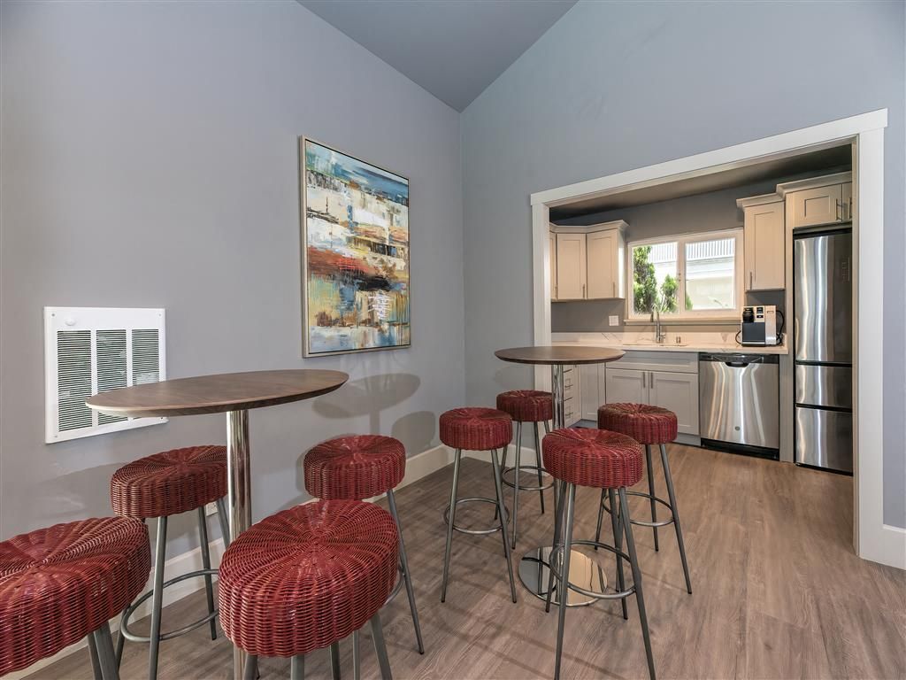 Common lounge with round wooden tables and red woven stools; kitchenette visible in background at Tower apartments in Alameda, CA.