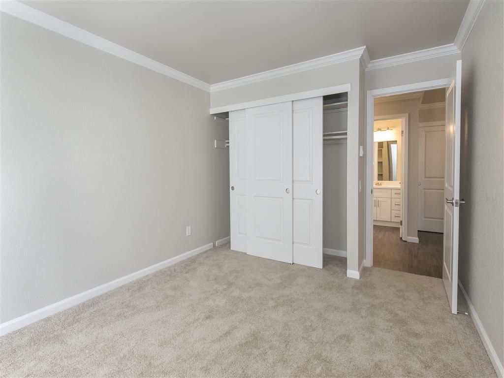Empty bedroom with beige carpet, white sliding closet doors, and an adjacent doorway to the bathroom at Tower apartments in Alameda, CA.