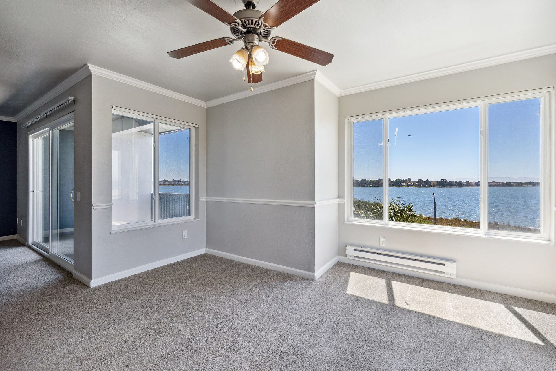 Bright living room with large waterfront view windows, ceiling fan, and sliding door to balcony at Tower apartments in Alameda, CA.