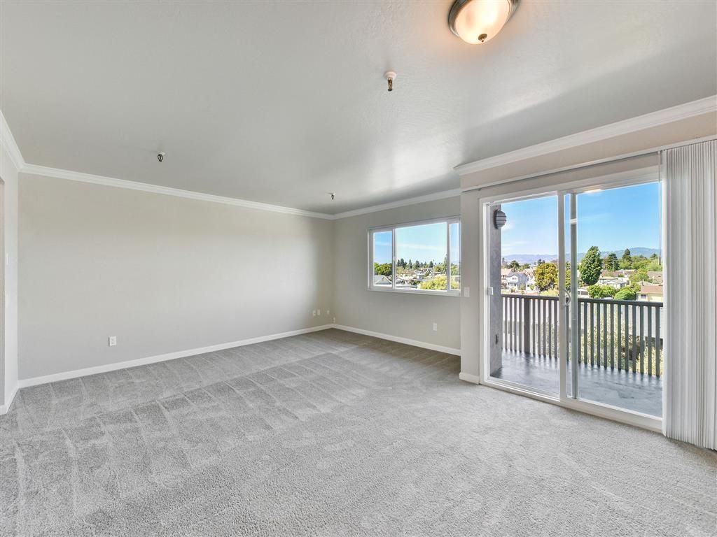 Bright empty living room with beige walls, gray carpet, and a sliding glass door to a balcony at Tower apartments in Alameda, CA.