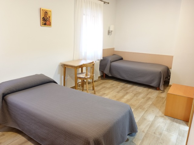Two beds in a simple room with a small table, chair, and wood-look floor.