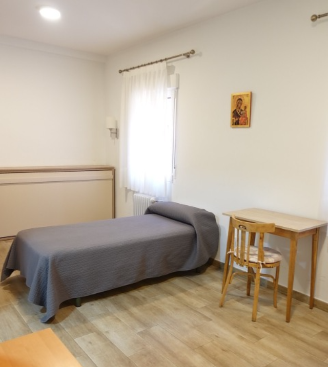 A simple bedroom with a bed, desk, chair, and small icon on the wall. Pale wood floor.