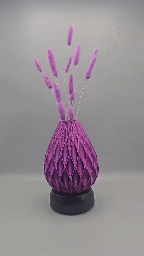 Een paarse vaas met een gestructureerd oppervlak en een traanvormig patroon staat op een zwarte voet en is gevuld met lange, vederachtige paarse stelen.