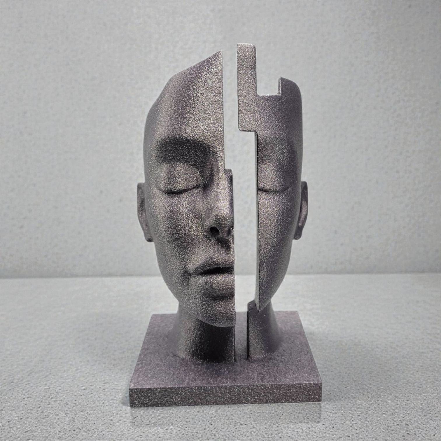 Een getextureerde, donkergrijze sculptuur van een menselijk gezicht, verticaal in twee delen gesplitst, geplaatst tegen een neutrale achtergrond.