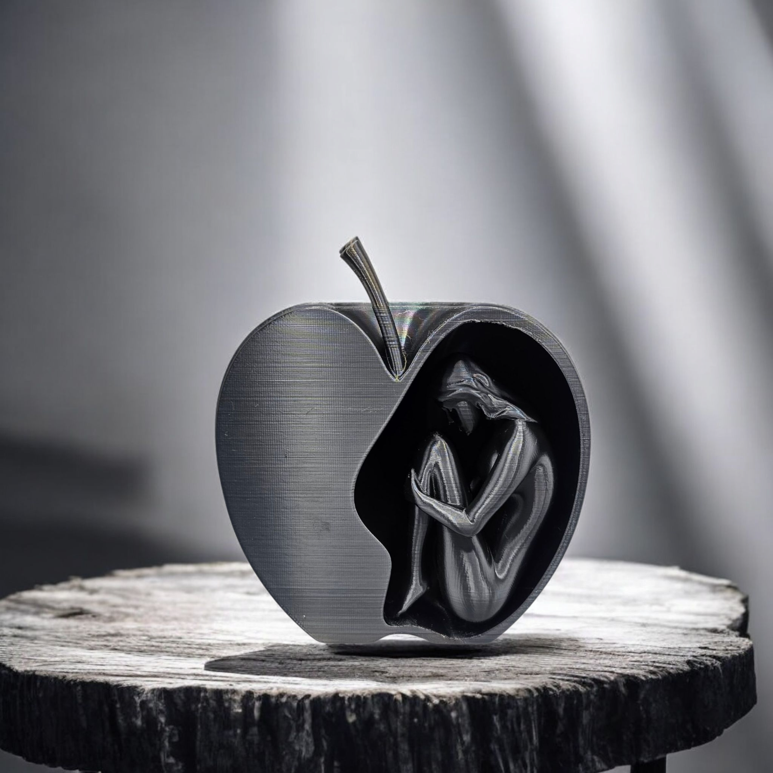 Een metalen sculptuur van een appel met een holle binnenkant waarin zich een klein figuurtje bevindt, opgerold in foetushouding, op een houten sokkel.