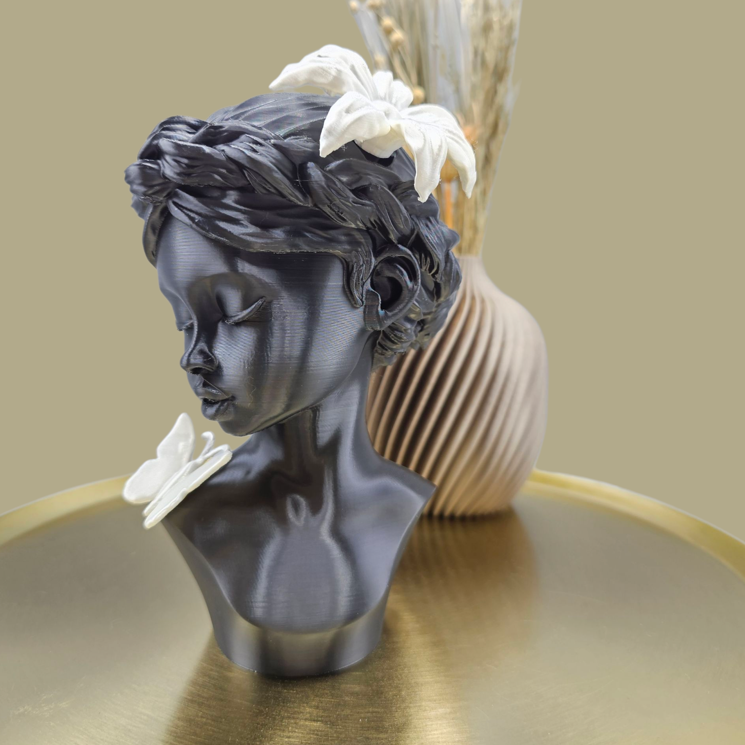 Een donkergrijze, 3D-geprinte buste van een persoon met een gevlochten kapsel, versierd met witte bloemen en een vlinder op één schouder.