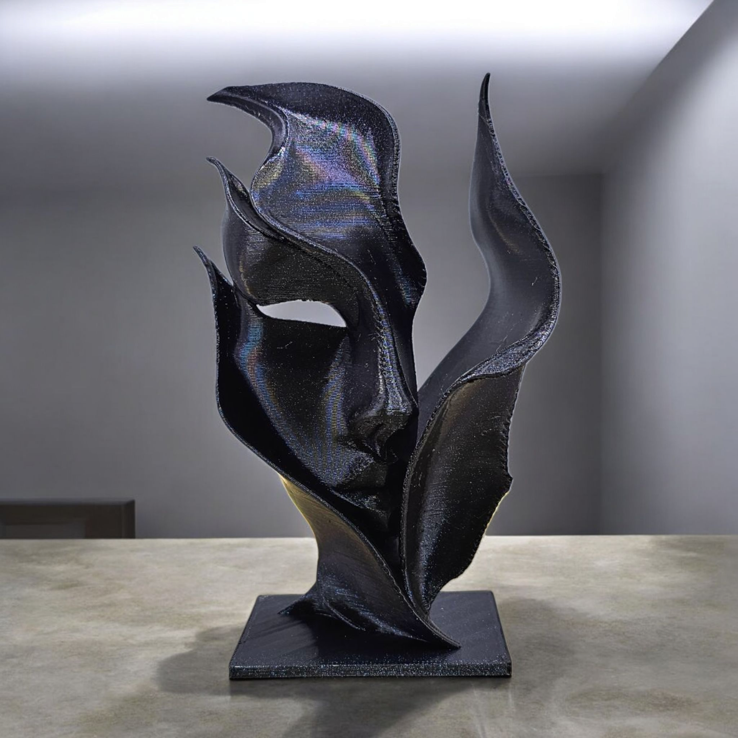 Een donkere, abstracte sculptuur met vloeiende, bloembladachtige vormen op een vierkante sokkel, geplaatst tegen een effen achtergrond.