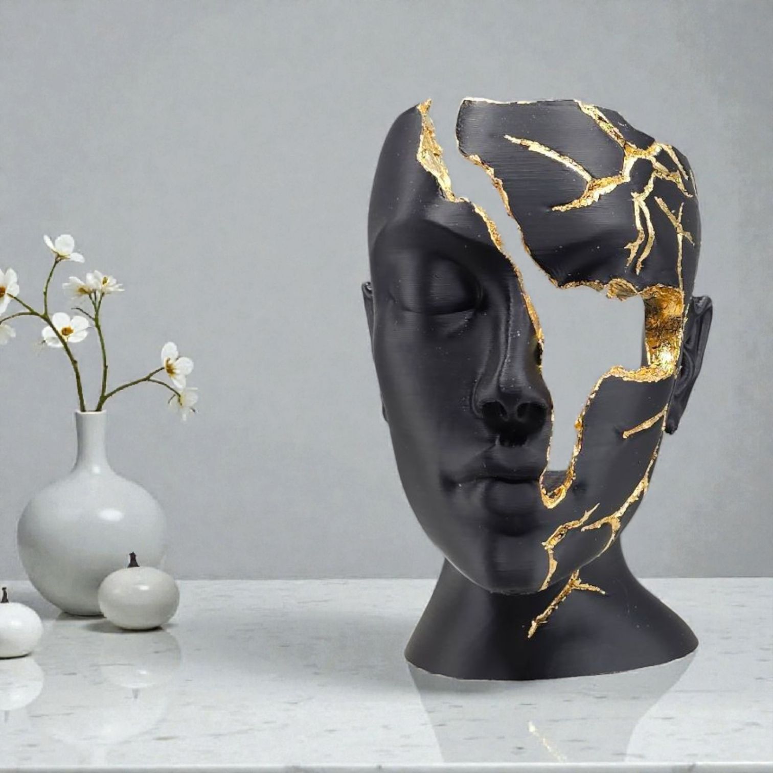 Een matzwarte sculptuur van een mensengezicht met accenten van bladgoud, die op een tafel een gebarsten en gebroken uitstraling heeft.