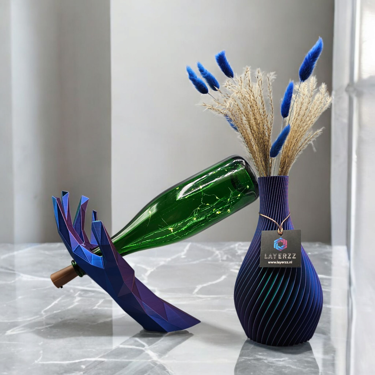 Een groene glazen fles staat in een geometrische, blauwe 3D-geprinte handhouder naast een gestructureerde, donkerblauwe vaas met gedroogde bloemen.