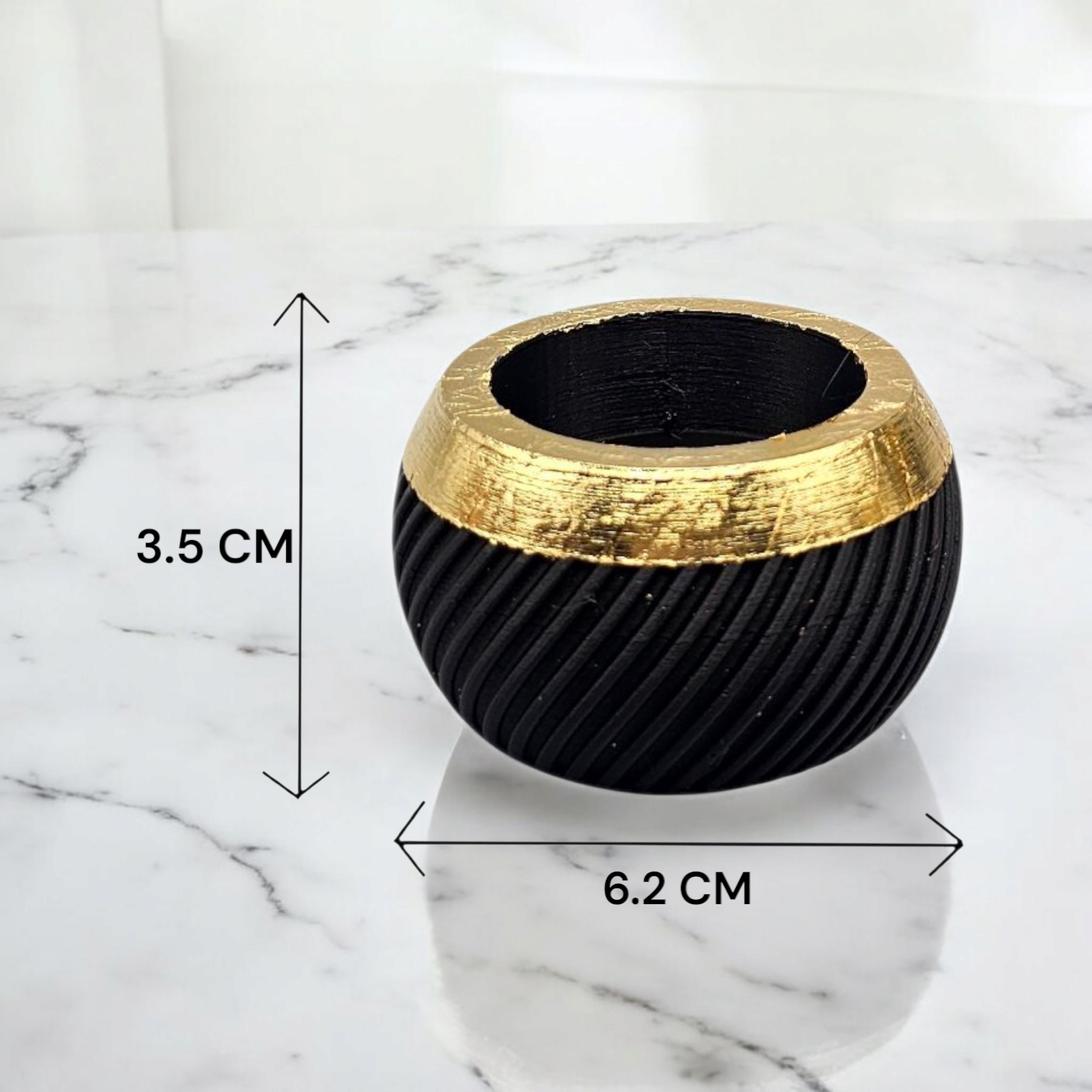 Een zwarte, gestructureerde, bolvormige kom met een gouden rand op een marmeren ondergrond. Hoogte: 3,5 cm, breedte: 6,2 cm.