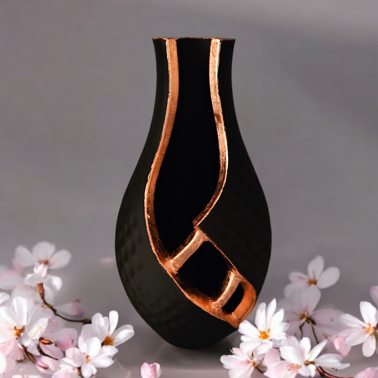 Een zwarte, druppelvormige vaas met een uitgesneden rand van metallic koper, omringd door roze bloemen op een grijze achtergrond.