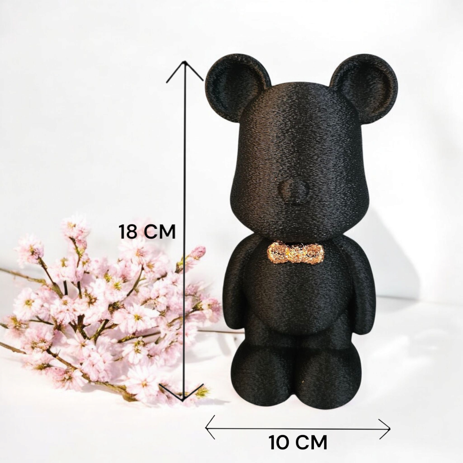 Een zwart, gestructureerd beeldje in de vorm van een beer met een klein gouden strikje, 18 cm hoog en 10 cm breed, naast roze bloemen.