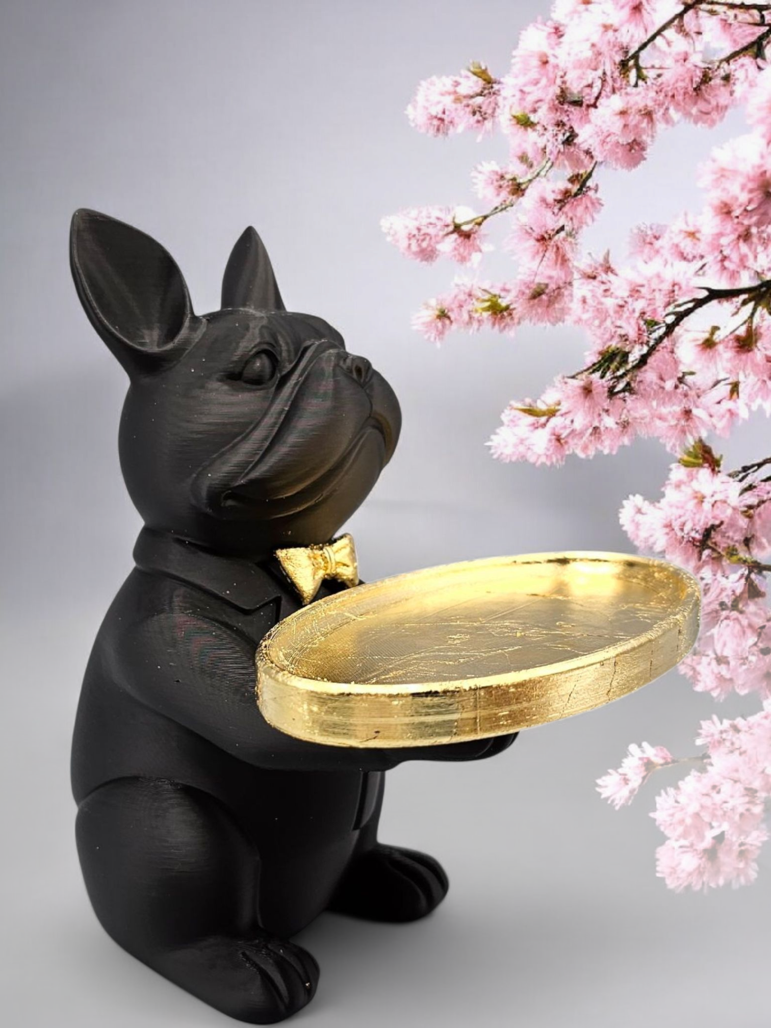 Een beeldje van een zwarte Franse bulldog met een gouden strik en een gouden dienblad, geplaatst tegen een achtergrond van kersenbloesems.