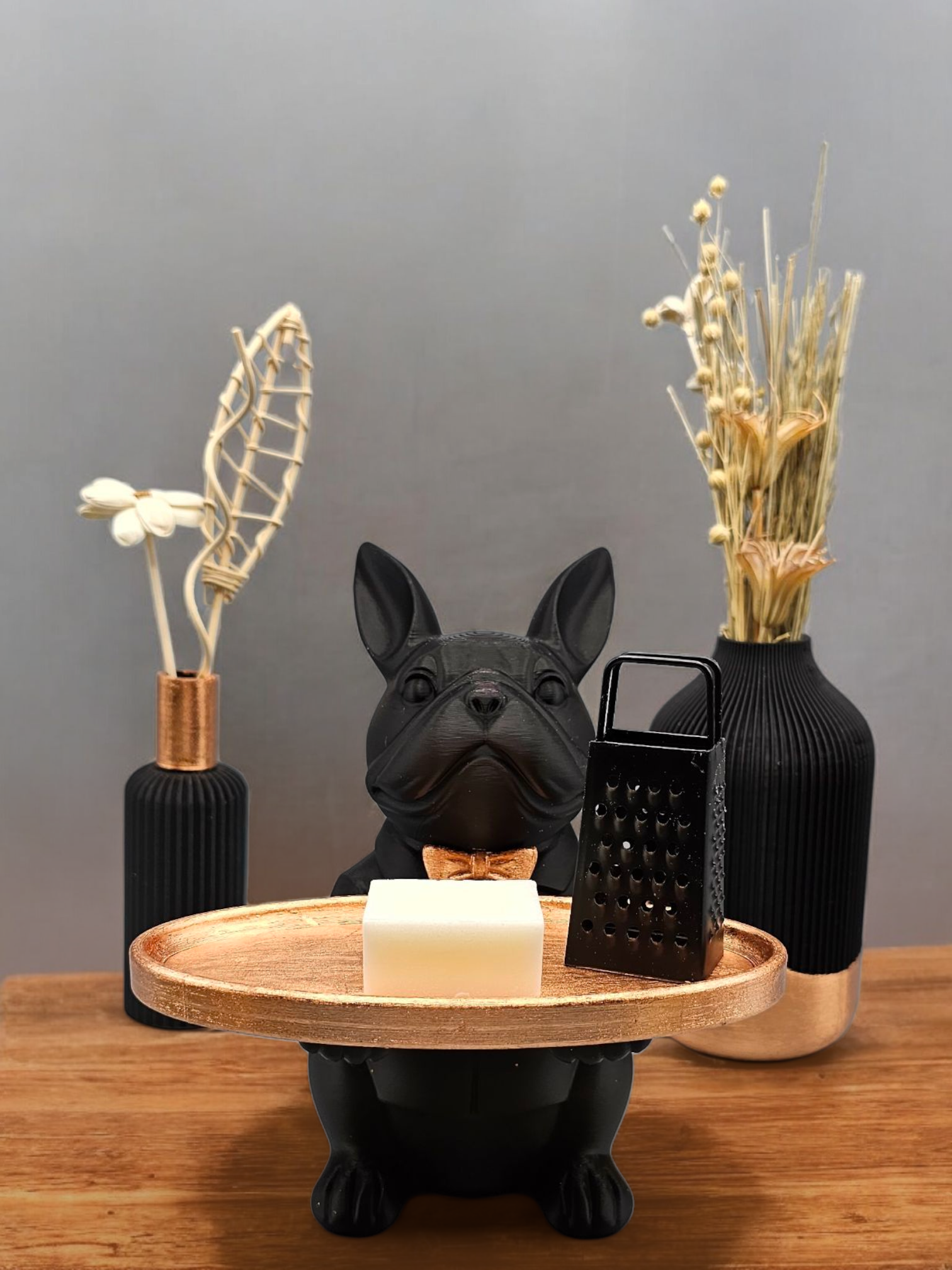Een beeldje van een zwarte bulldog dat een gouden dienblad vasthoudt met een wit blokje en een kleine rasp, geflankeerd door twee vazen met gedroogde bloemen.
