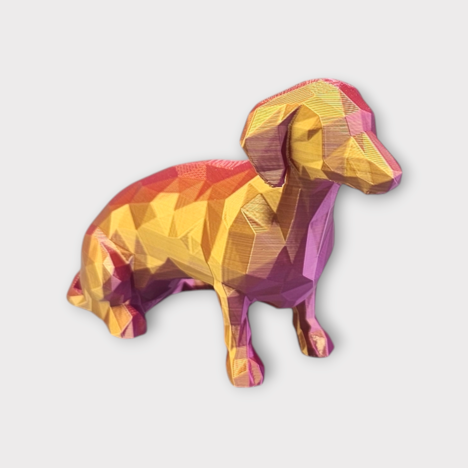 Een low-poly, 3D-geprint teckelfiguurtje met een levendige, metallic kleurovergang van goud, oranje en magenta.