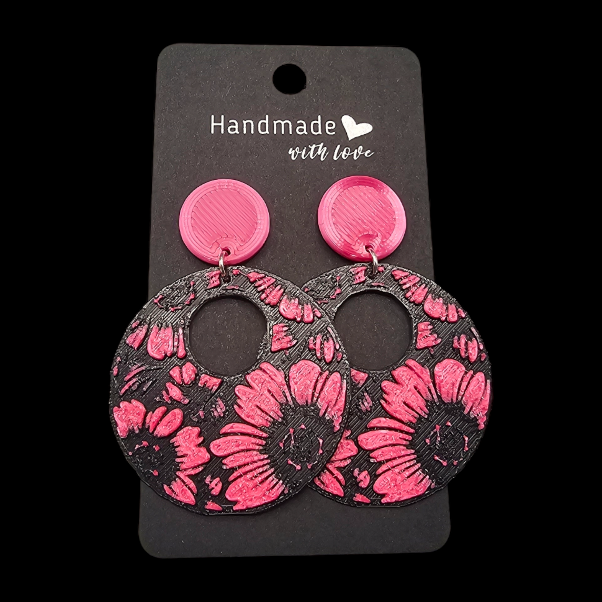 Handgemaakte oorbellen op een presentatiekaartje met ronde, felroze oorstekers en grote, ronde, zwarte hangers met een roze bloemenprint.