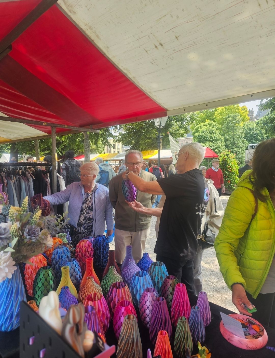 Op een openluchtmarkt toont een verkoper een kleurrijke vaas met een bijzondere textuur aan klanten achter een tafel vol soortgelijke artikelen.
