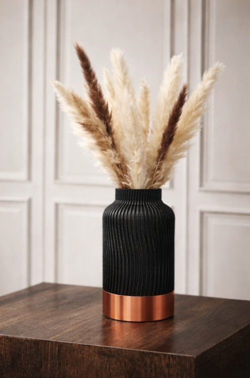 Een zwarte, gestructureerde vaas met een koperen voet staat op een houten ondergrond, gevuld met beige en bruin pampasgras.