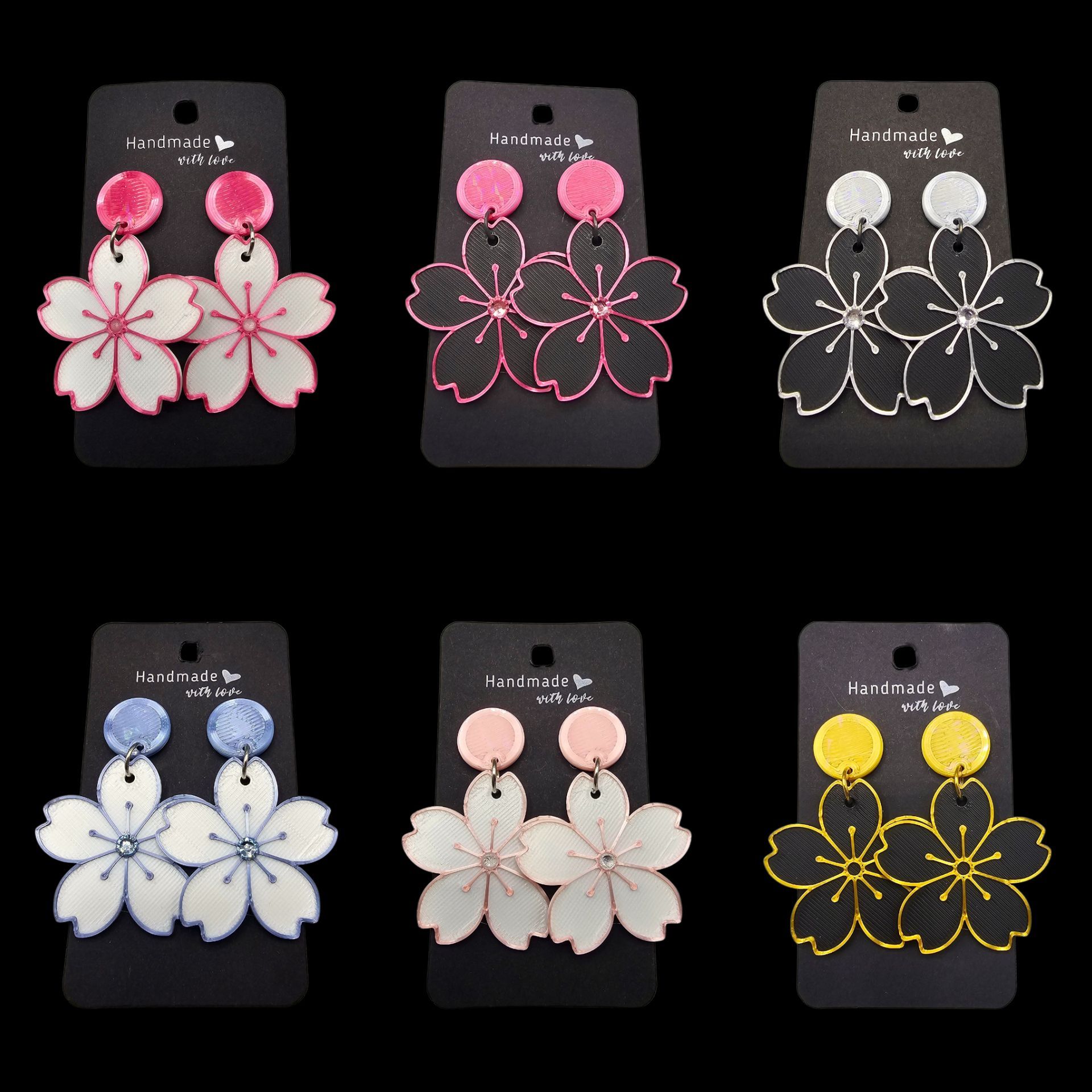 Zes paar oorbellen in de vorm van kersenbloesems op zwarte presentatiekaartjes, met diverse kleurencombinaties van roze, zwart, blauw en geel.
