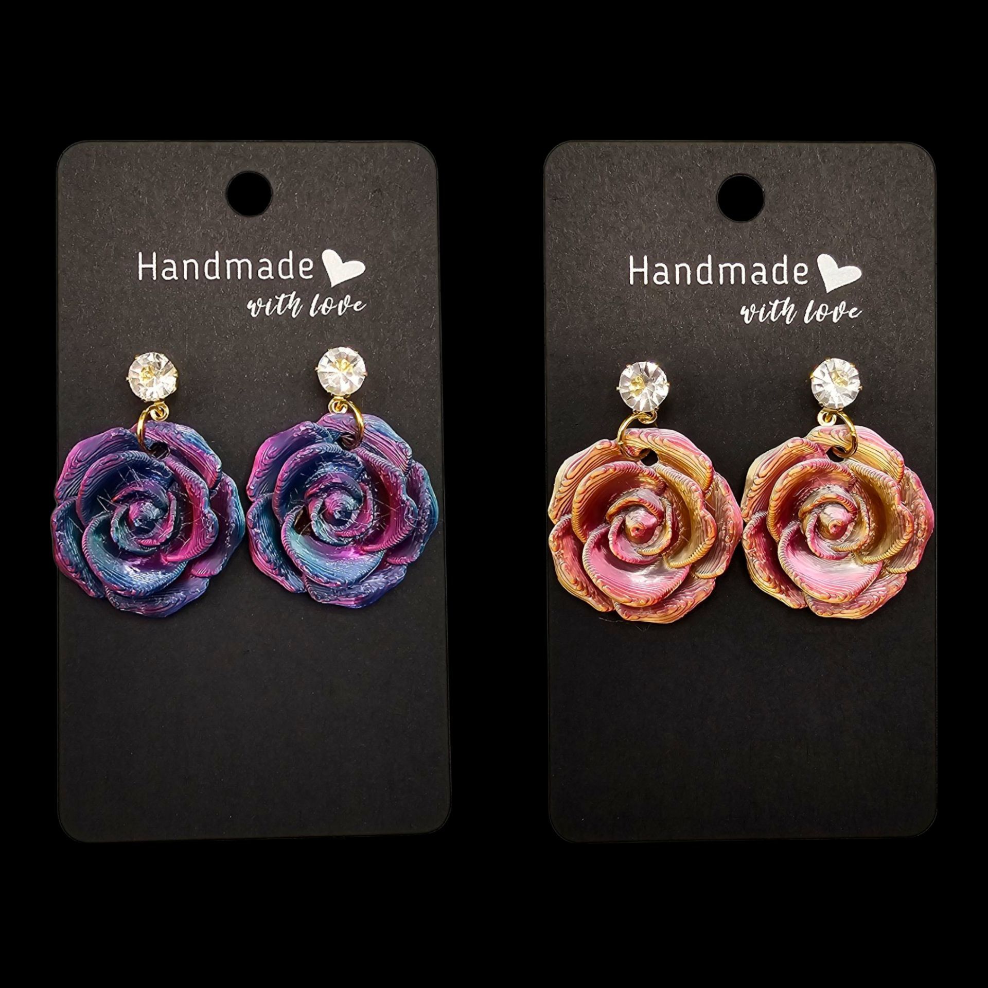 Twee paar handgemaakte rozenoorbellen met strasssteentjes op zwarte presentatiekaartjes: een paarsblauwe set met een galaxy-motief en een roze-gouden set.