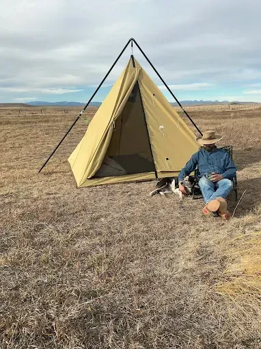 A person in a hat and a dog sit in chairs outside a tan pyramid-style tent in a vast, dry field.