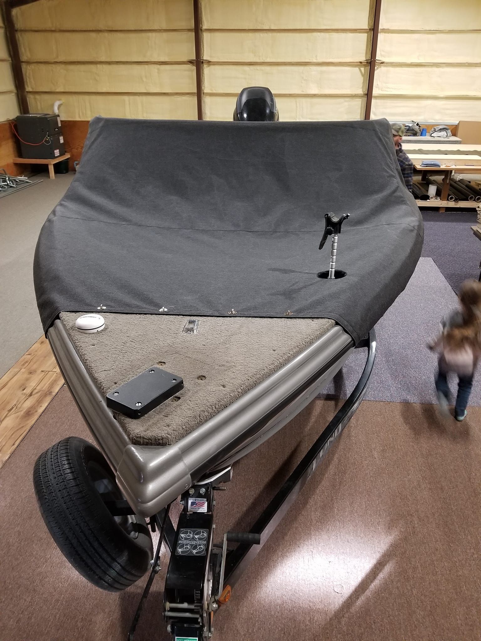 A boat on a trailer, covered with a gray tarp, parked in a garage with a blurry figure visible to the right.