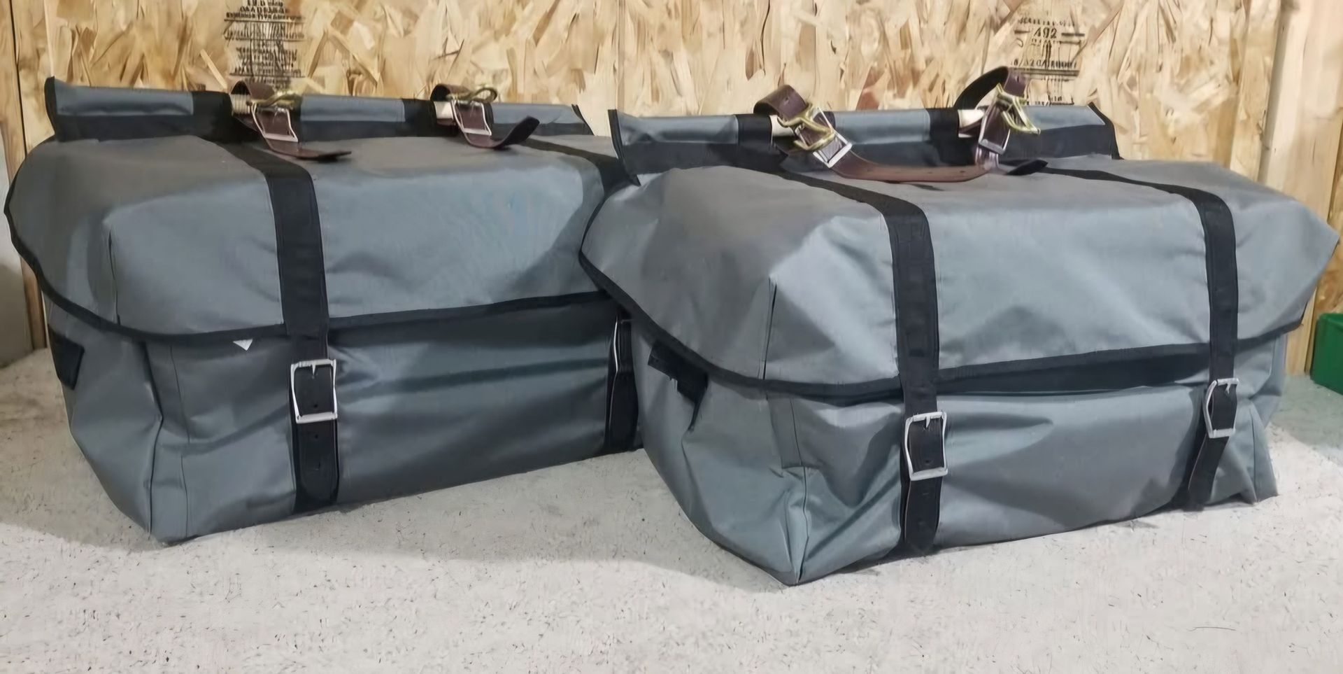 Two gray rectangular storage bags with black straps, buckles, and brown leather accents sitting on a light surface.