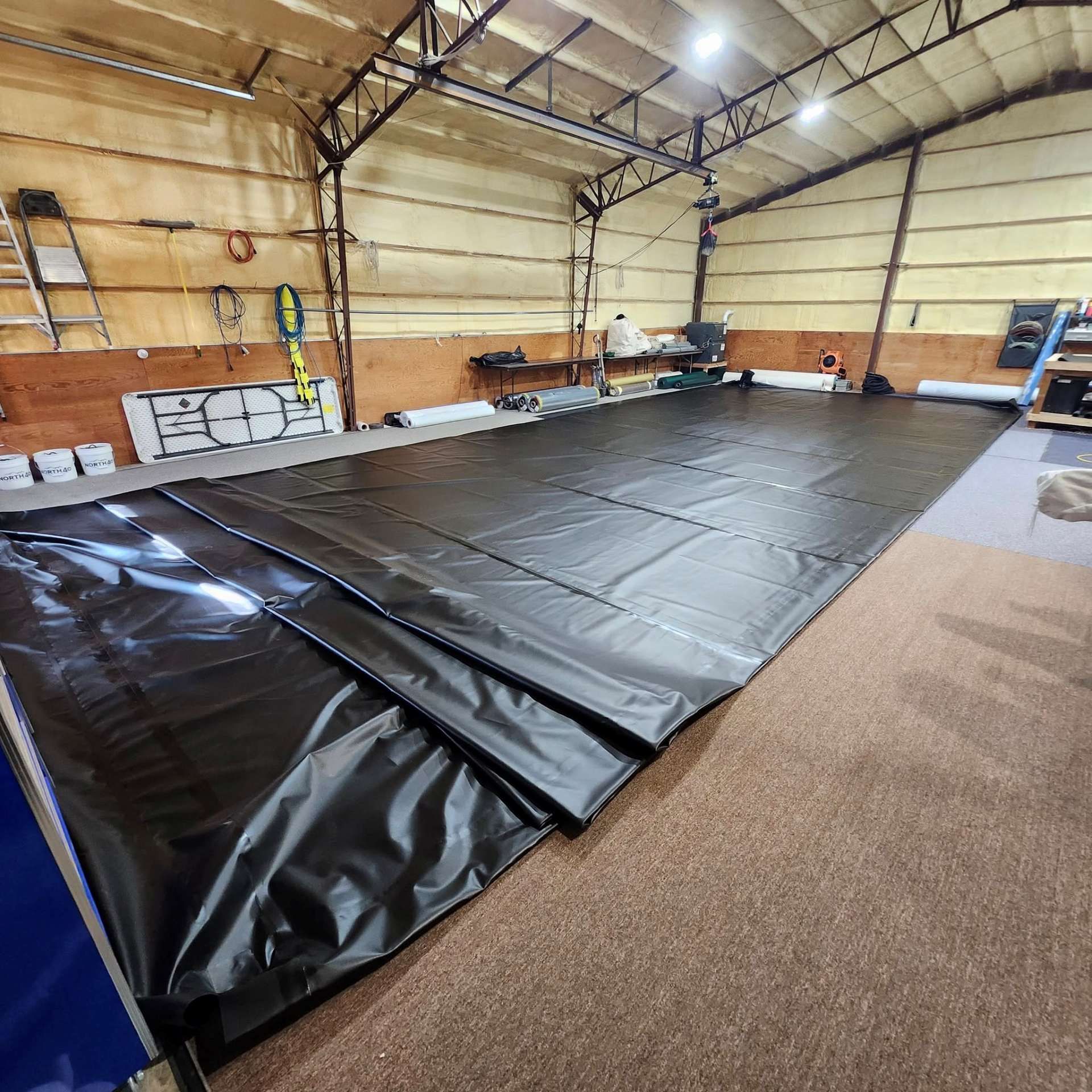 A large black tarp or liner covers the floor of a spacious workshop with high, wood-paneled walls and exposed rafters.
