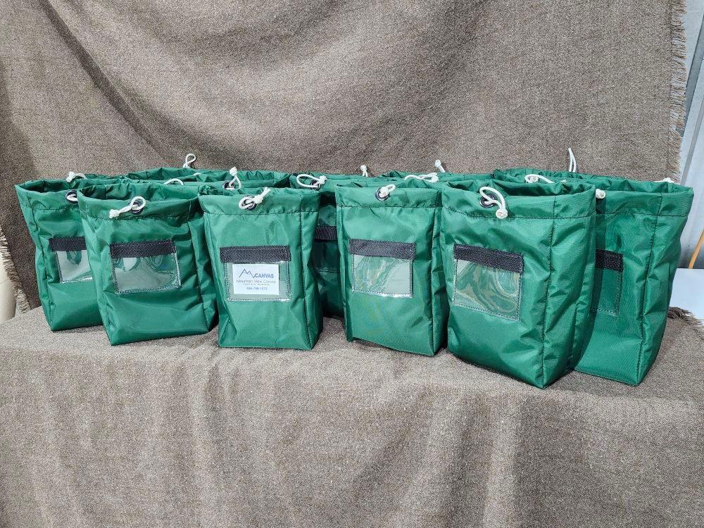 A row of seven green drawstring pouches with clear rectangular display windows, sitting on a burlap-covered surface.