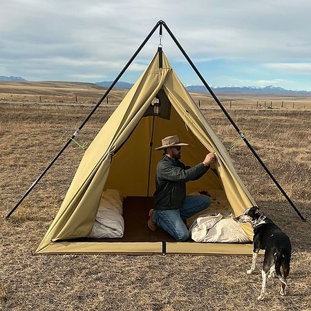 A person wearing a hat sits inside a tan, pyramid-shaped canvas tent in a grassy field, next to a dog.