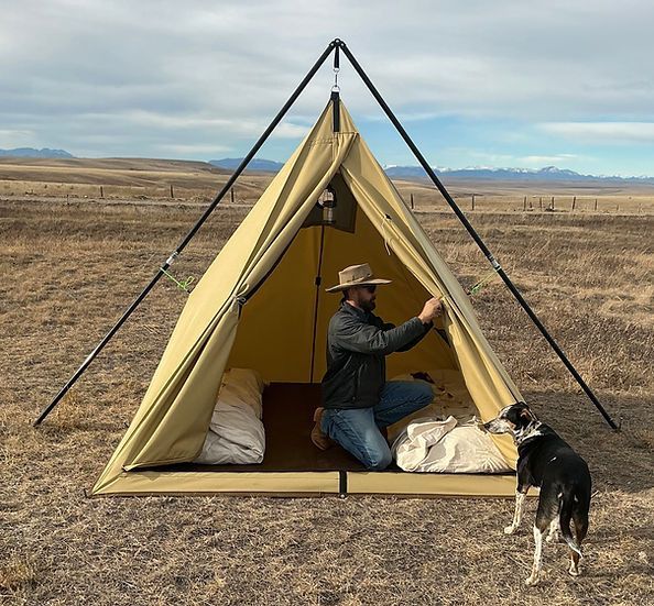 A person in a cowboy hat sets up a tan teepee-style tent in a desert landscape with a dog nearby.
