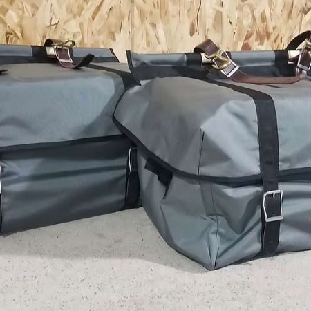 Two grey, rectangular, weather-resistant storage bags with black webbing straps and metal buckles, set against a wood wall.
