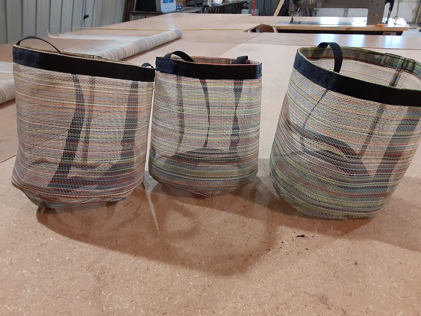 Three woven mesh baskets with dark, solid-colored rims sit in a row on a wooden workshop surface.