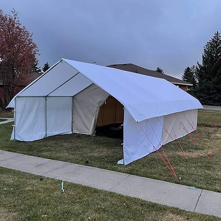 A large, white canvas wall tent with a metal frame set up on a residential lawn next to a sidewalk.