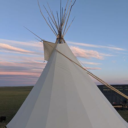 A white canvas teepee stands in a grassy field against a sky with sunset clouds.