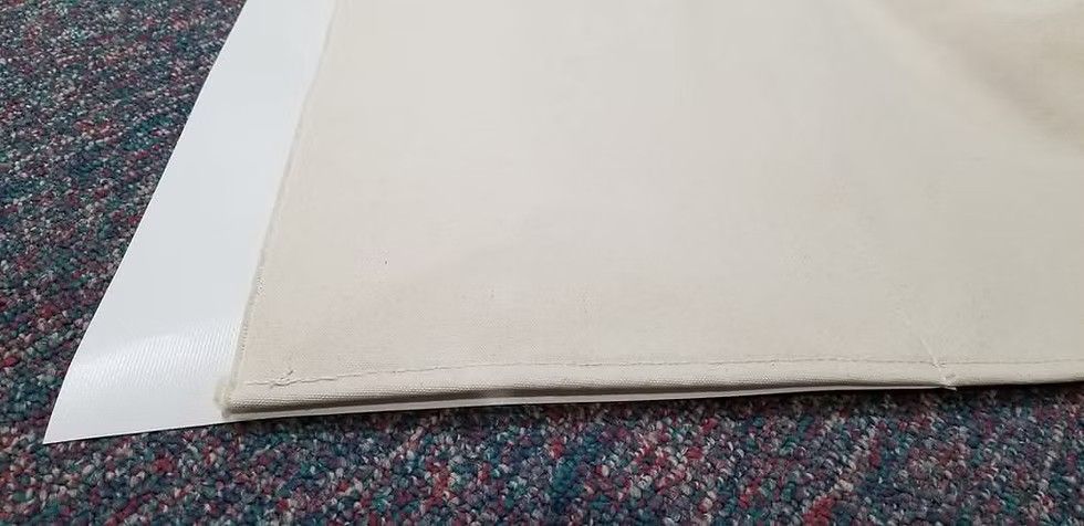 A beige fabric edge with a hemmed seam resting partially over a smooth white surface on a dark carpeted floor.